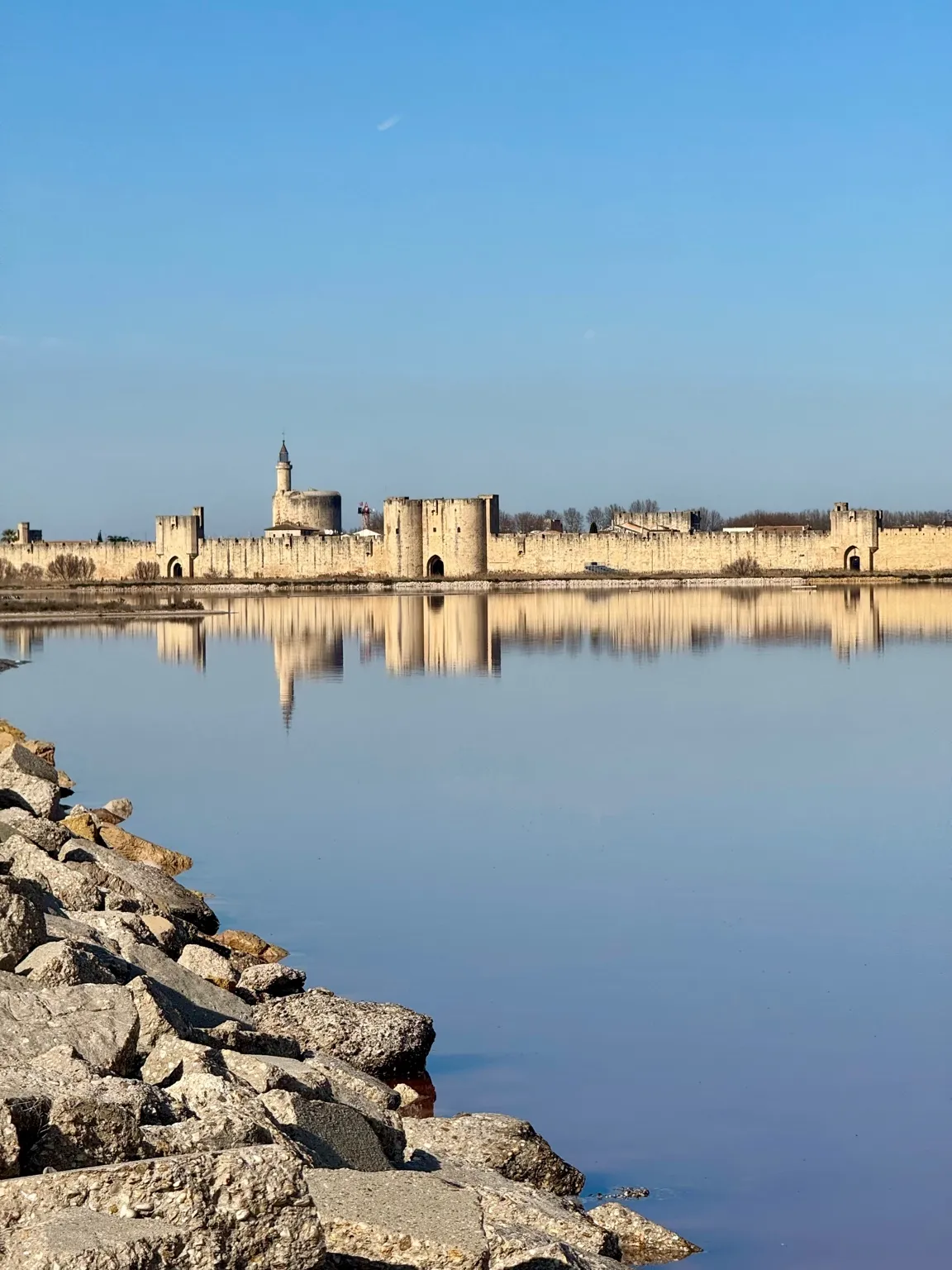Odbicie średniowiecznych murów Aigues-Mortes w spokojnej wodzie, pod bezchmurnym niebem.
