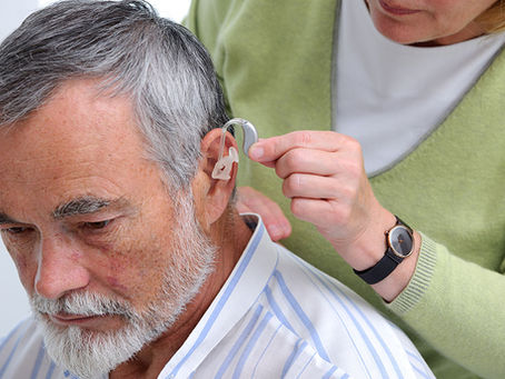 An elderly man with gray hair receives assistance with his hearing aid from a person in a green sweater. The setting is calm and focused.