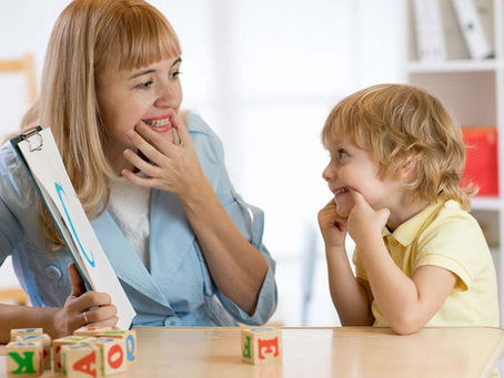 A woman with a clipboard and a smiling child imitate facial expressions. Colorful alphabet blocks are on the table in a bright room.