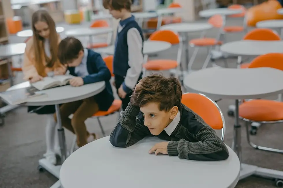 Boy with hand on chin sits at a round table in a library, looking pensive. Three kids read a book nearby. Orange chairs are around.