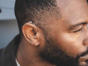 Close-up side profile of a bearded man wearing a silver hearing aid, looking down against a soft gray background.