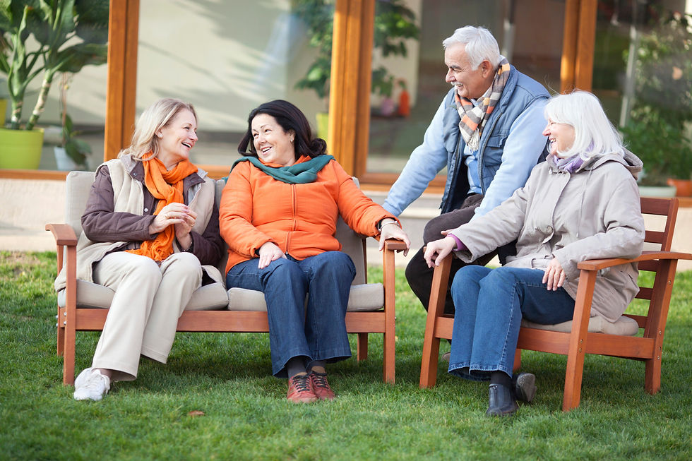 Four seniors sit and chat happily on wooden chairs in a garden. They're wearing colorful jackets and scarves, creating a warm, cheerful scene.