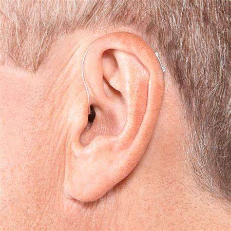 Close-up of an ear with a small, beige RIC hearing aid. The skin is light-toned, and the background is out of focus, emphasizing the device.