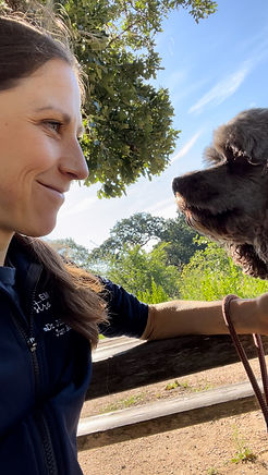 A cisgendered woman with brown hair smiling at a cockapoo dog as they sit outside in the sun.