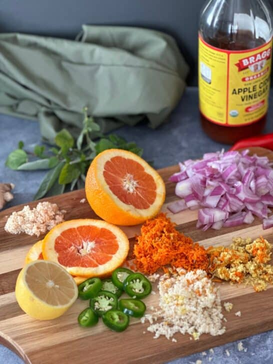Sliced citrus, onions, and spices on a wooden board with a bottle of apple cider vinegar. Green cloth and herbs in the background.