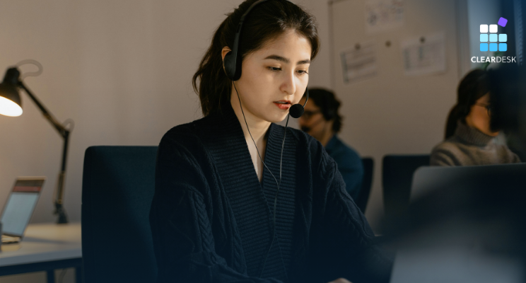 Woman in a black top wearing a headset, working intently at a computer in an office. Logo in corner says CLEAR DESK. Cozy lighting.