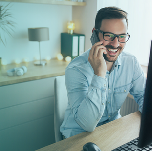 A man holding a phone, smiling while talking, seated at a desk with a desktop computer in a bright workspace.
