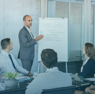 A man standing and presenting annual goals to his team, speaking confidently while team members sit around the table, actively listening.