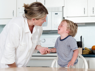 Woman in white shirt comforts a crying child in a kitchen. White cabinets and a blue countertop with fruit in the background.