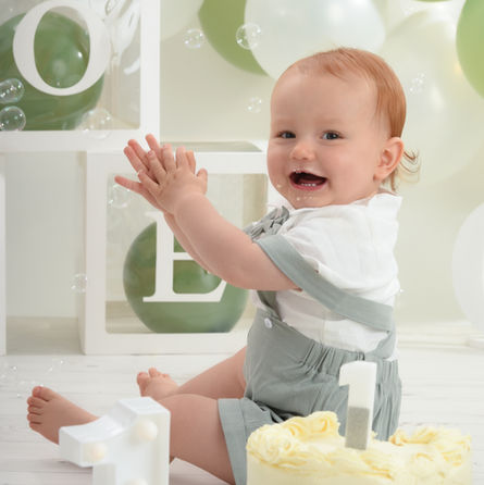 Baby boy sat by his cake smash birthday cake, clapping his hands and trying to catch bubbles in the air