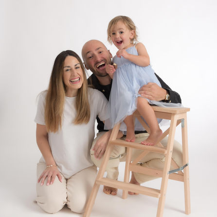 Mum dad and little girl smiling and happy. Little girl is wearing a blue party dress and sitting on a ladder.