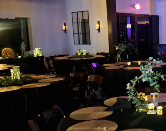 Dimly lit banquet hall with round black tables, tan placemats, candles, greenery, and purple accent lighting.