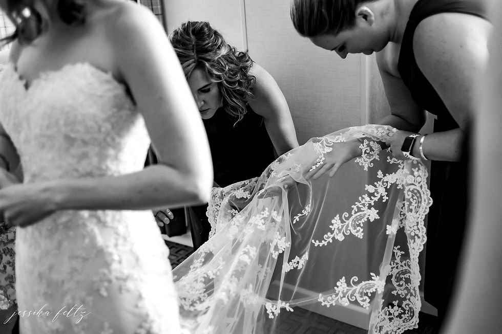 Black-and-white wedding prep: bridesmaids adjust a lace veil on a bride in a dressing room; watermark reads jessika feltz