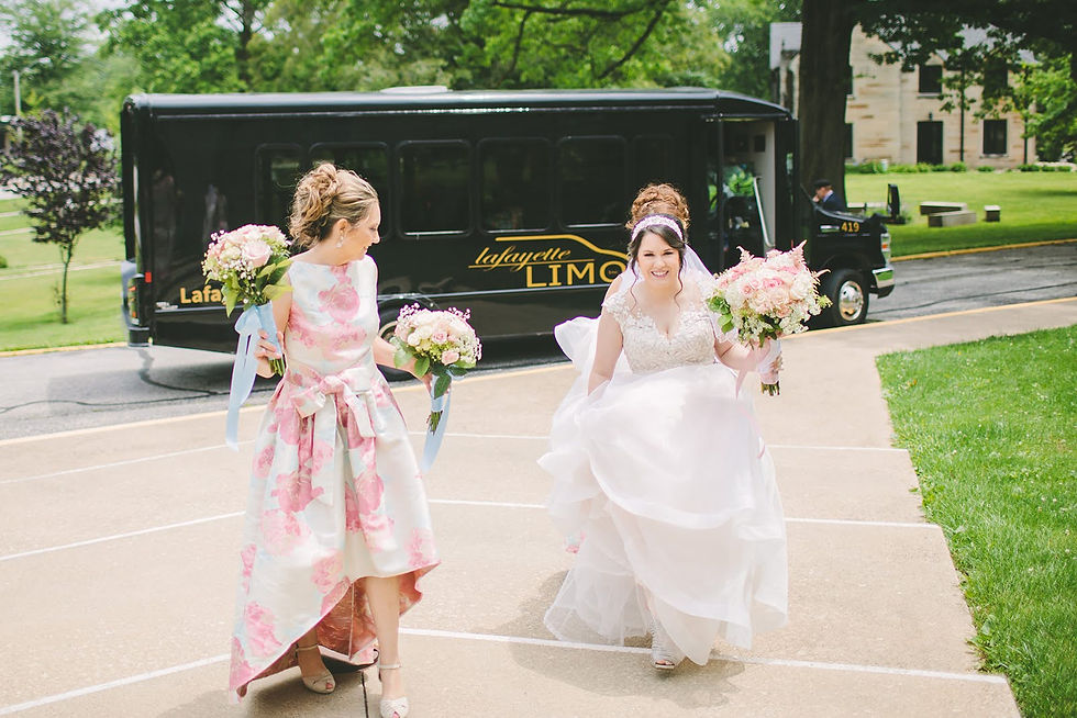 Bride and woman in floral dress hold bouquets, smiling, walking on a path. A black limo with "Lafayette Limo" visible in the background.