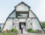 Bride and groom kiss in front of a white barn, surrounded by greenery and gardens, on a sunny day. Romantic and serene setting.