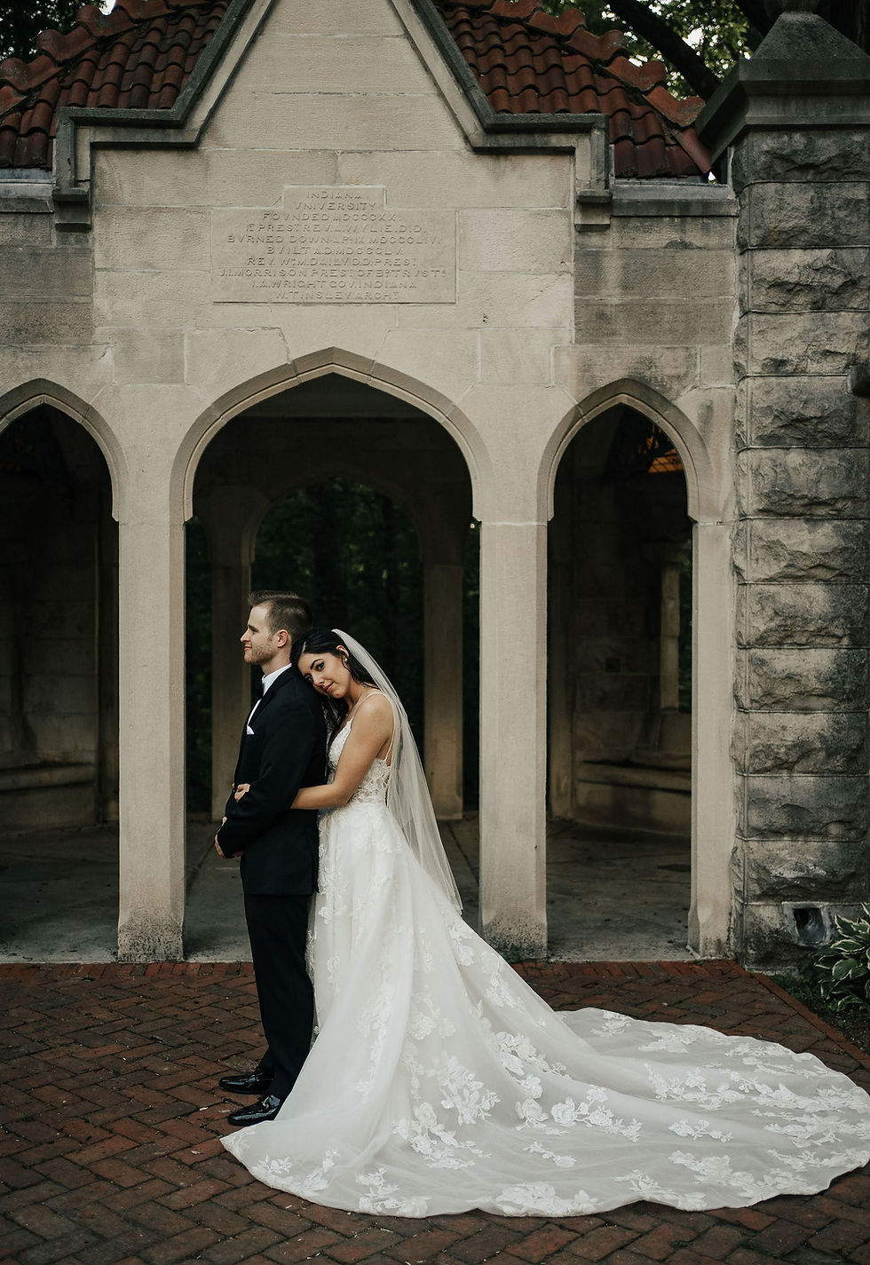 Bride in white gown hugs groom in black tuxedo beneath a stone archway with an Indiana University plaque.