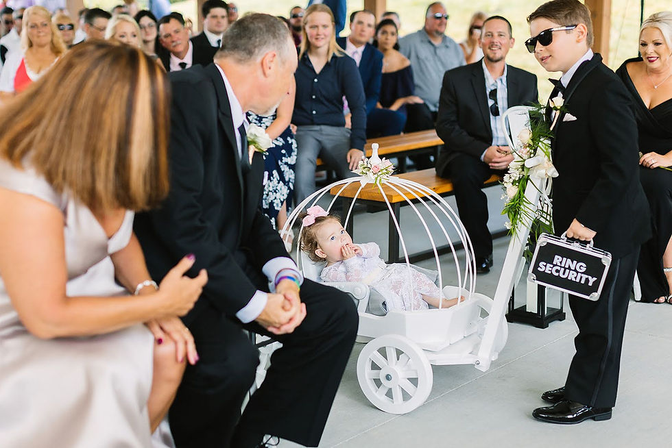 Wedding guests watch a boy in tux and sunglasses holding a Ring Security case beside a toddler in a white cart with flowers