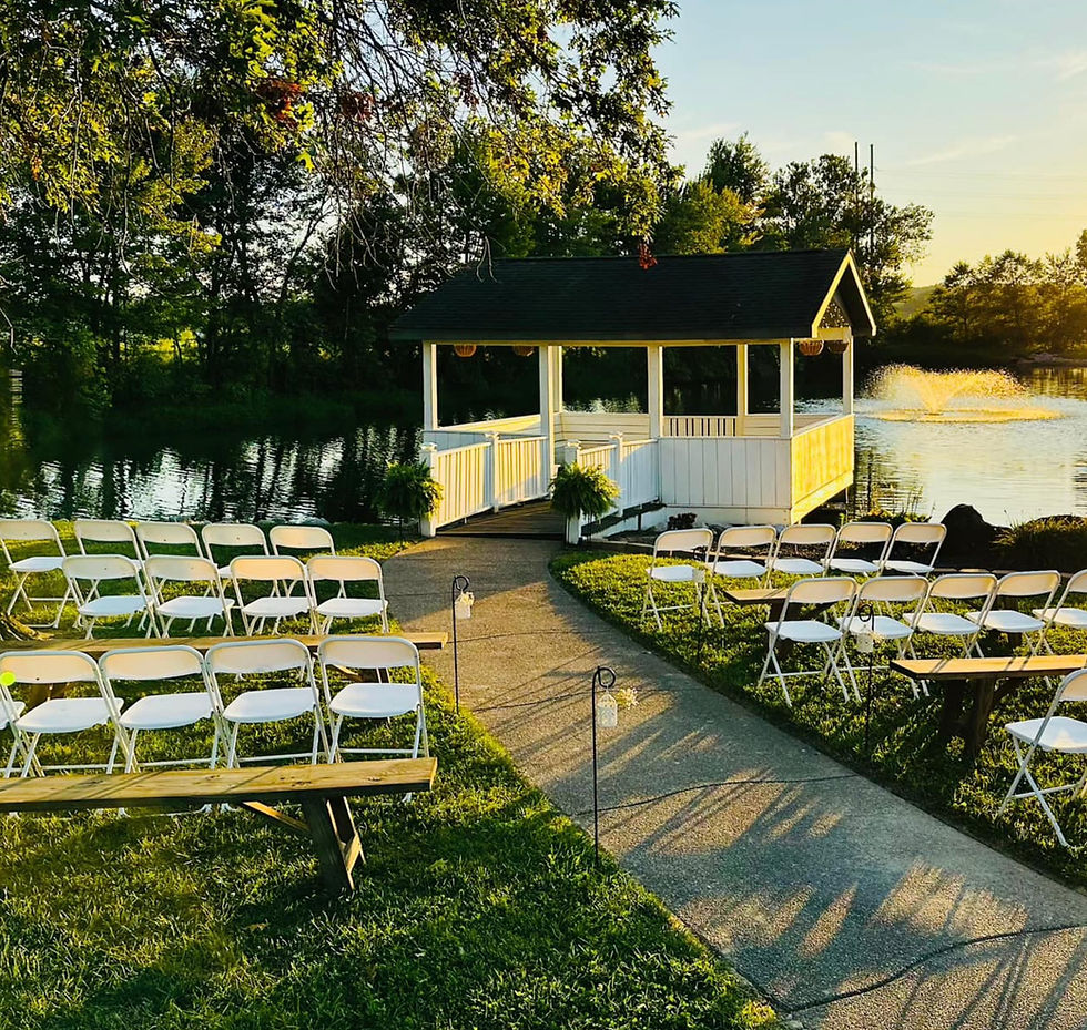 Outdoor lakeside wedding setup with white chairs and benches leading to a gazebo, golden sunset light, and a fountain beyond.