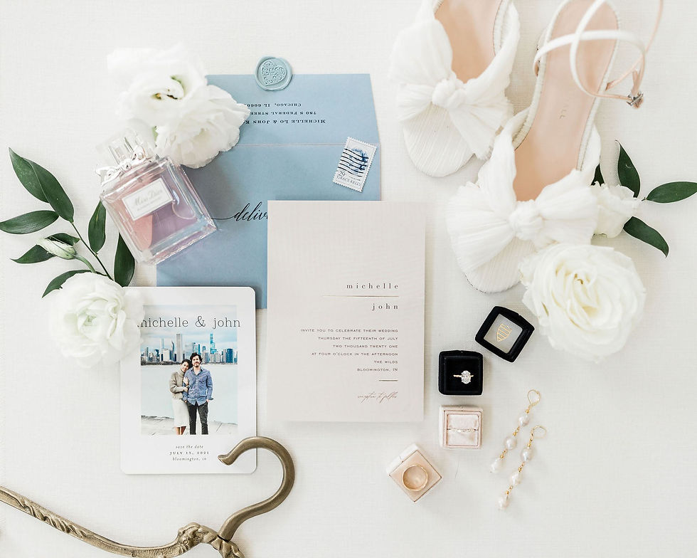Flat lay of wedding invitations, white floral heels, perfume, flowers, rings, and jewelry on a soft ivory background.