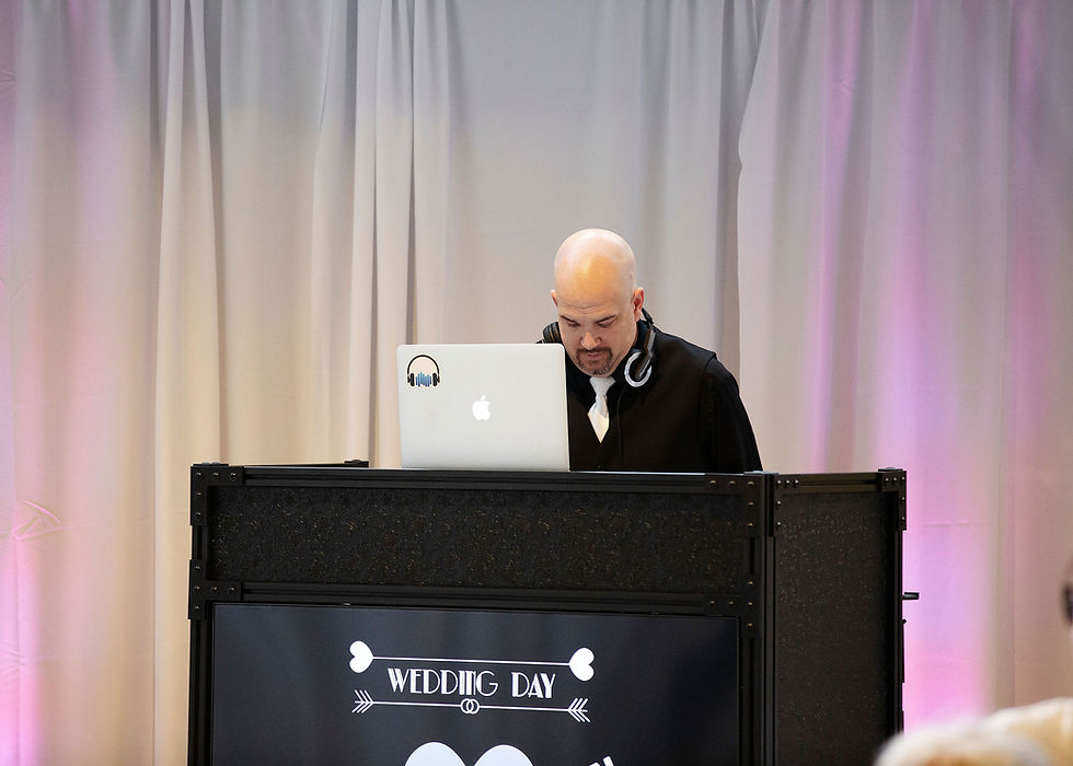 DJ in black suit with headphones behind a laptop, in front of a Wedding Day sign and soft pink-lit curtains.