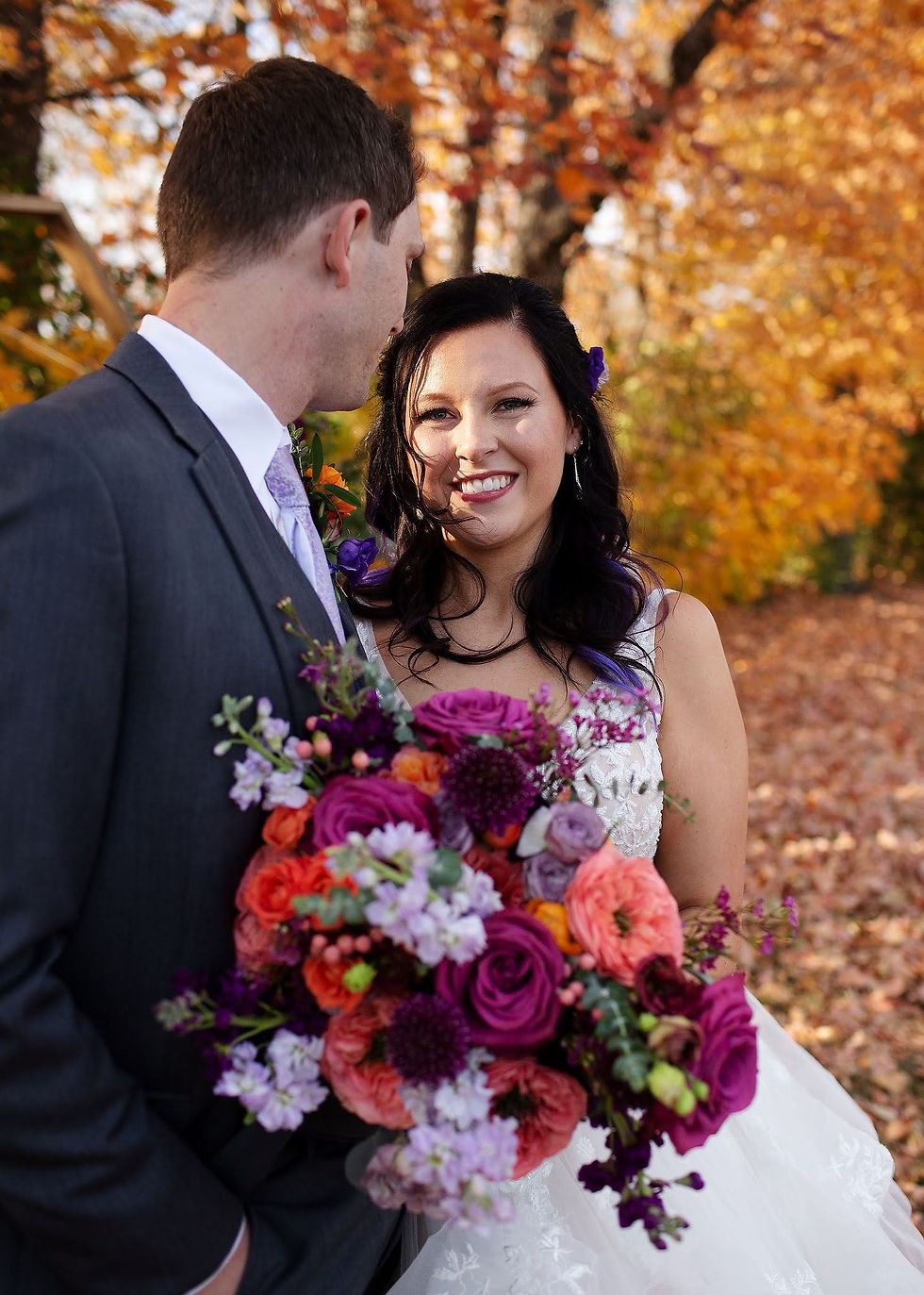 Bride smiling at groom in suit, holding vibrant bouquet with purple and orange flowers. Autumn trees with golden leaves in background.