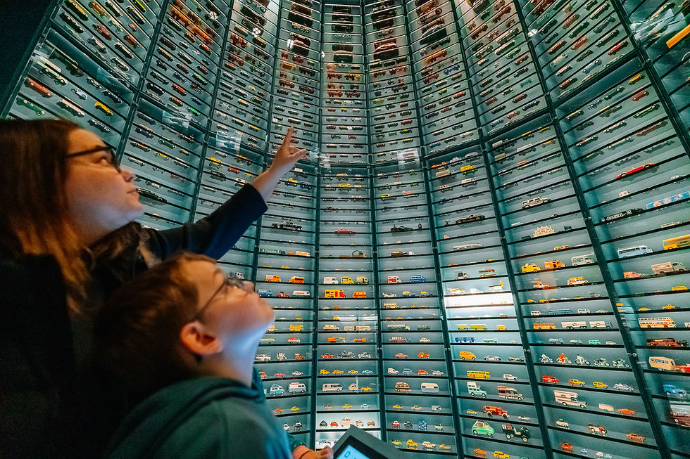 Zwei Kinder zeigen in einem Museum auf eine hohe Glaswand voller Modellautos; staunende Stimmung.