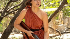 Ana de Armas in Eden (2025), dressed in a one-shoulder rust-red outfit and headscarf, holding a shotgun while standing in a sunlit, rugged island setting.