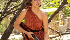 Ana de Armas in Eden (2025), dressed in a one-shoulder rust-red outfit and headscarf, holding a shotgun while standing in a sunlit, rugged island setting.