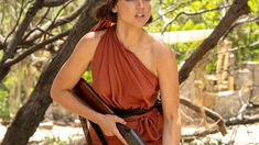 Ana de Armas in Eden (2025), dressed in a one-shoulder rust-red outfit and headscarf, holding a shotgun while standing in a sunlit, rugged island setting.