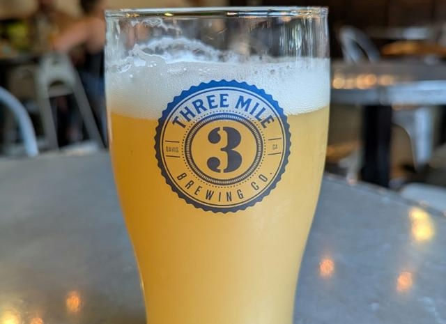 A beer in a beer glass with the three mile logo on it sitting on a table outside in the sunshine.