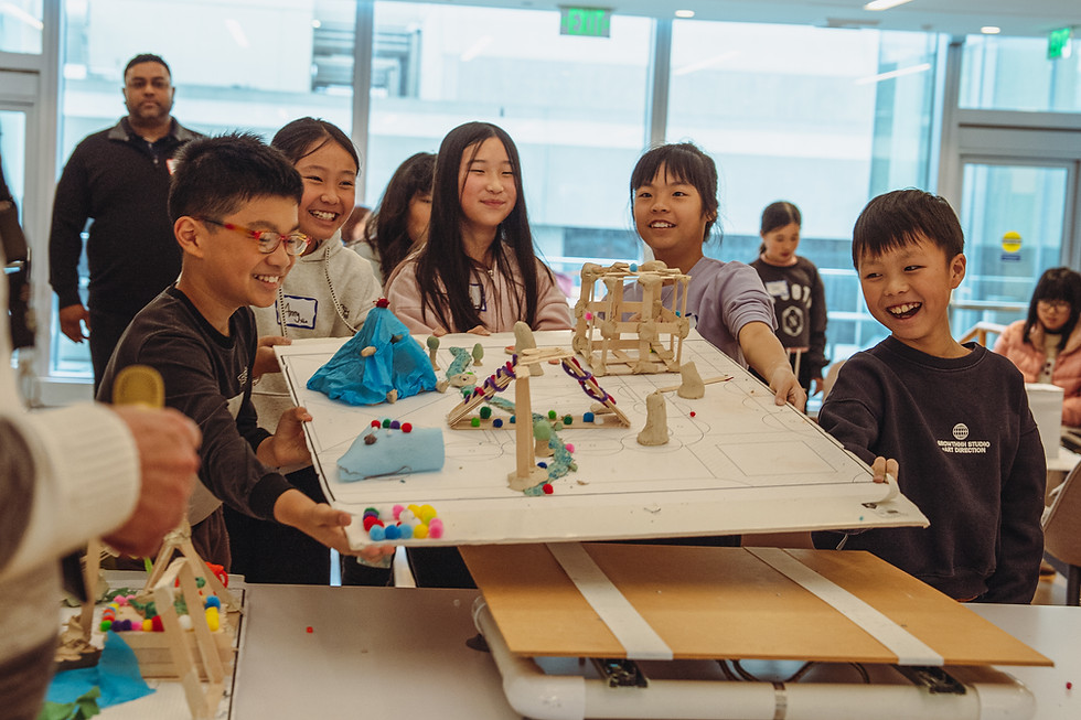 A group of young children carrying their project that they built of a small town made of kids arts materials to a table where its structural integrity will be tested by shaking.
