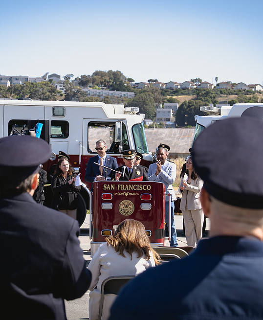 San Francisco Fire Department Chief Dean Crispen speaks to a crowd in front of a backdrop of firefighters and fire trucks.