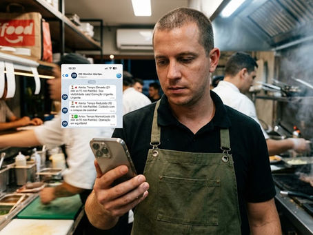Uma fotografia profissional em estilo 'candid' de um homem, que aparenta ser o gerente ou proprietário de um restaurante, em pé dentro de uma cozinha comercial movimentada. Ele veste uma camiseta escura e um avental de lona verde, e tem uma expressão focada enquanto olha para a tela de um smartphone segurado em suas mãos. A tela do telefone está brilhantemente iluminada, exibindo um aplicativo de chat com várias mensagens e notificações em português, incluindo textos claros sobre alertas operacionais como 'Alerta: Tempo Elevado' e 'Alerta: Tempo Reduzido', indicando que ele está revisando um sistema de monitoramento de entregas. O fundo, levemente desfocado, mostra o ambiente agitado de uma cozinha industrial moderna, com outros funcionários trabalhando e uma sacola de entrega vermelha com o logo 'iFood' visível em uma prateleira, fornecendo contexto.