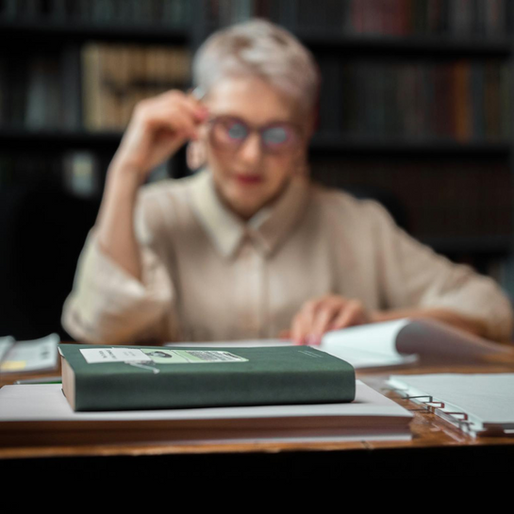 A closed green book with a white label and an open book, with a person with short white hair and glasses in the blurred background, sitting at a wooden desk in a library, reading.