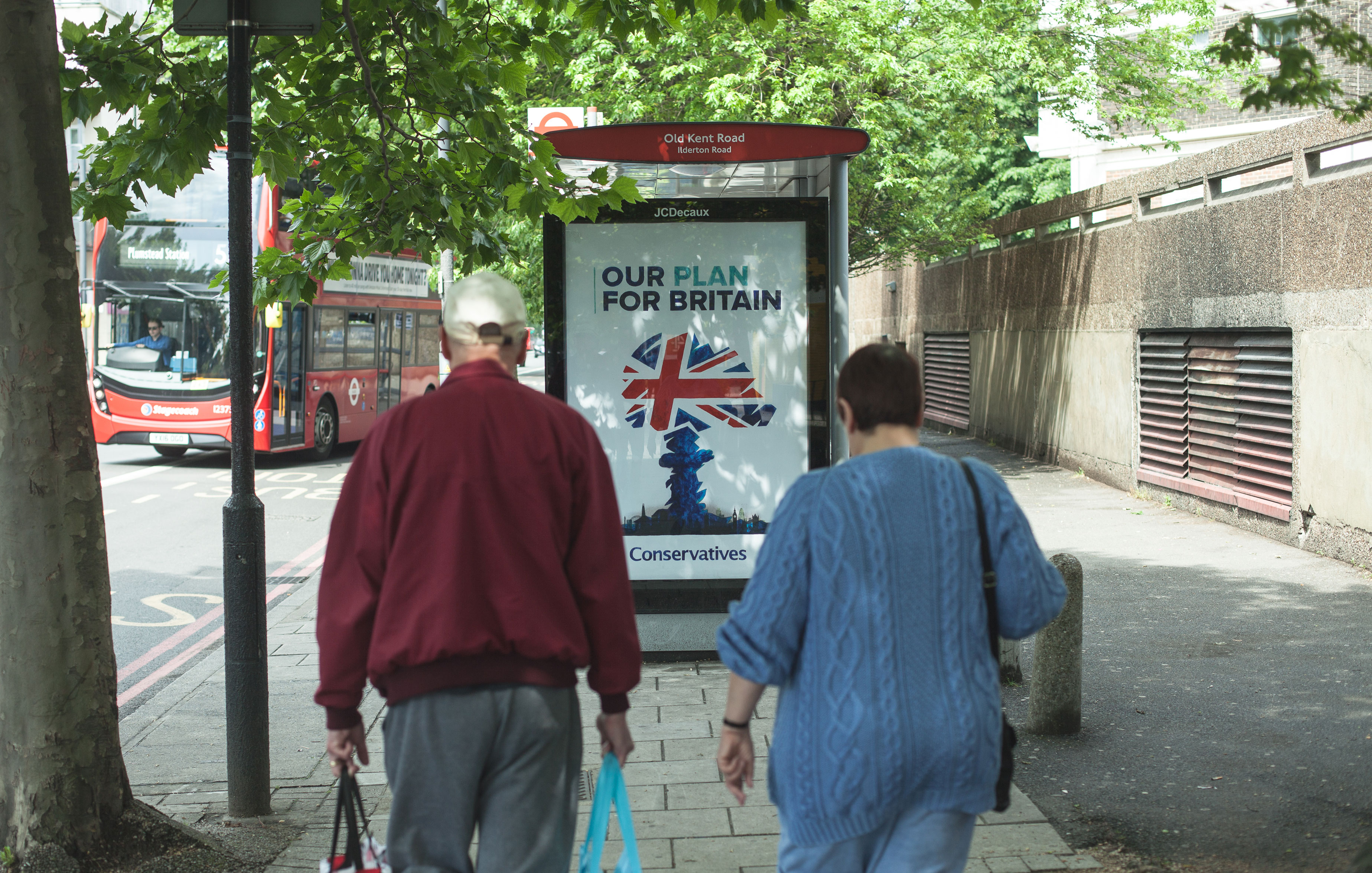 Two people walking towards an anti-Tory subvertising ad hacked bus stop poster. The poster is the Conservative party logo redone as a nuclear mushroom cloud