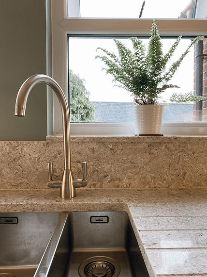 a kitchen window with a plant on the sill. Stone worktops and a stainless steel sink and tap 