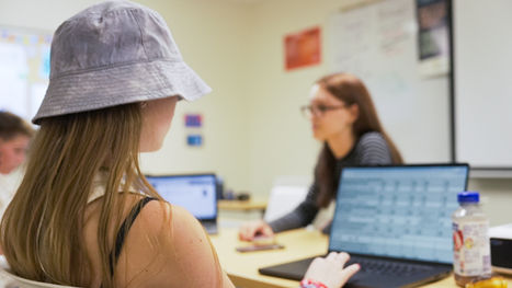 Student in a gray bucket hat uses a laptop in a classroom while a blurred teacher looks on; other laptops and a drink bottle nearby