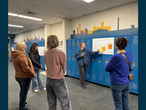 Communities for Youth staff member points at a bar graph poster on the wall, presenting to a group of four people in a hallway with blue lockers.