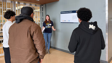 Communities for Youth staff member presents a poster titled  "Youth Substance Use by Category" to a group of three students. 