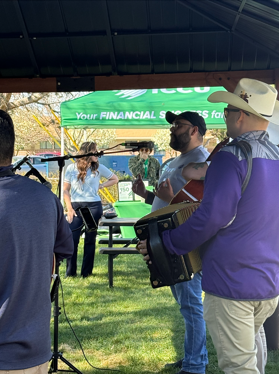 Musicians holding a guitar and accordion listen to Alex Zamora speak into a microphone under a pavilion