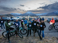 Cyclists and spectators gather in a mountainous parking area. People smile and give thumbs up. Snow-capped peaks in background.
