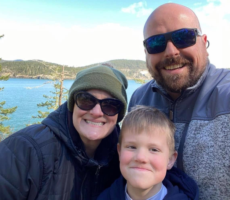 Smiling family selfie of two adults and a young boy posing by a blue lake with forested hills.