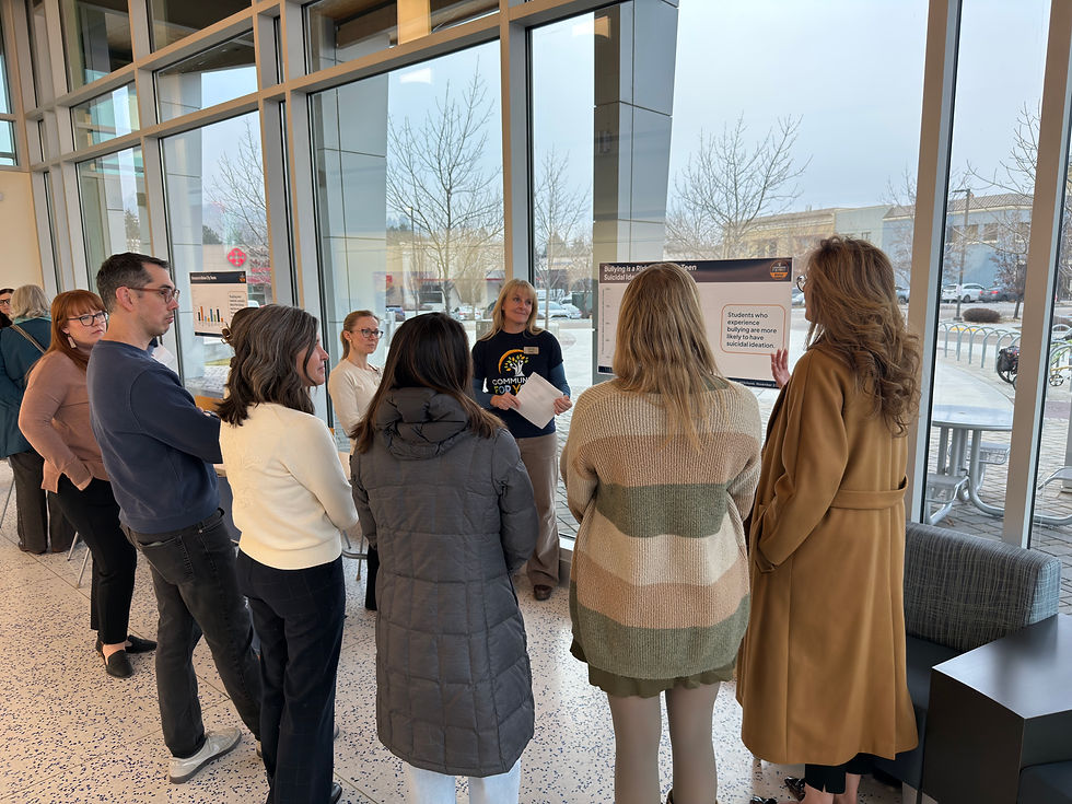 Small group listens to a presenter beside posters about bullying and suicide risk in a bright glass-walled lobby