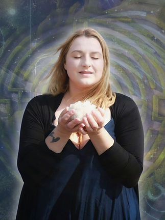 A female holding a crystal cluster in her hands while smiling softly. In the background is a labyrinth.