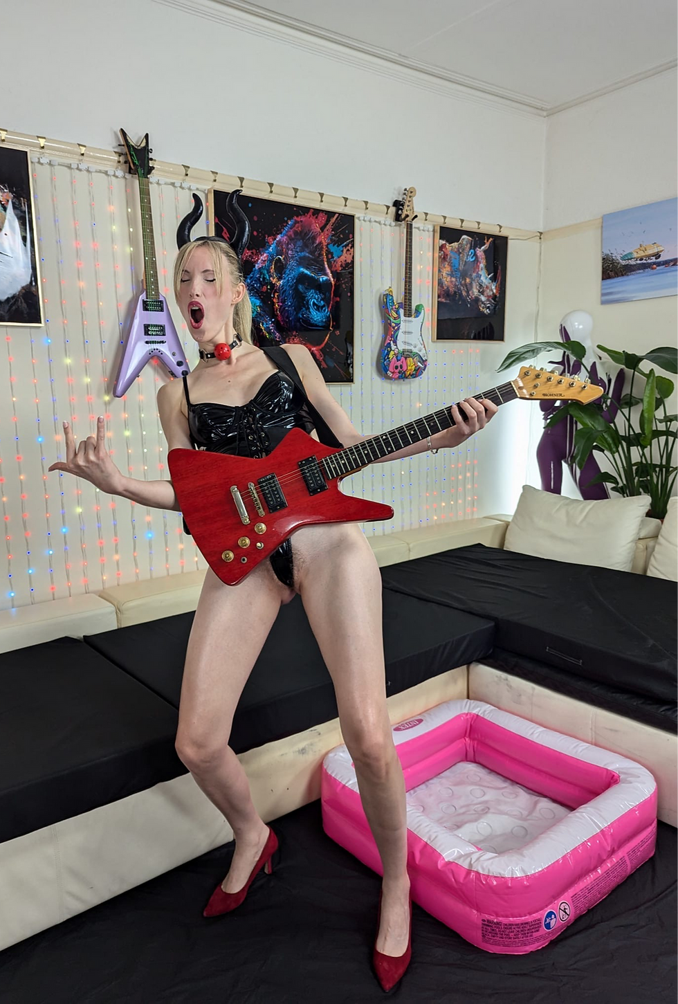 Nausi Love holding a red electric guitar in a living room beside a pink inflatable pool.