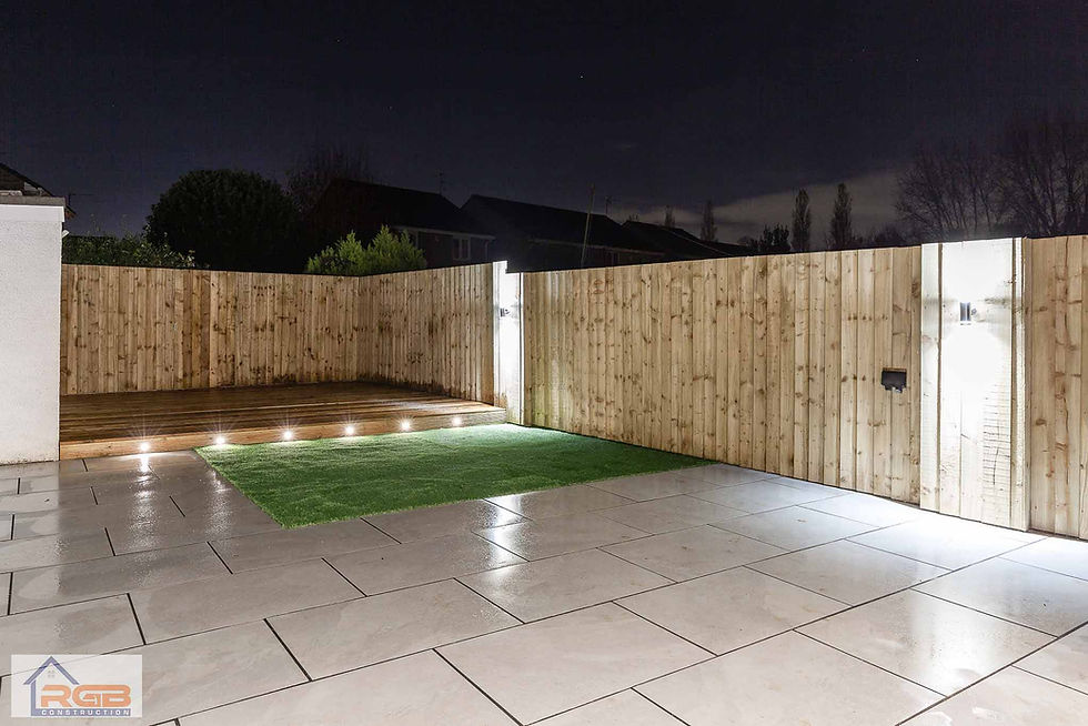 Completed modern landscaped garden featuring a paved patio, artificial turf, timber decking, and ambient outdoor lighting at night.