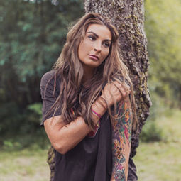Fashion portrait of female alternative model with arm tattoos wearing black ACDC t-shirt while leaning against a tree posing with her hand on her shoulder in a forest at the Craig Yr Hesg Nature Reserve in South Wales