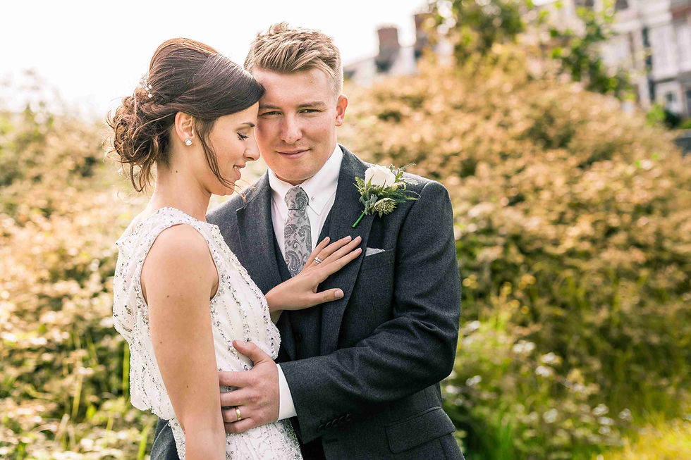 Groom looking at camera while holding his bride as she admires her wedding ring with her hand on his chest in a field outdoors