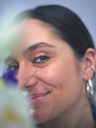 Junge Frau mit zurückgebundenem Haar lächelt hinter unscharfen Blüten, Farbportrait
