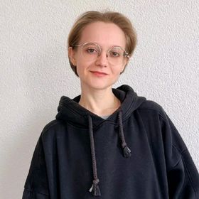 Person with short hair and glasses smiles, wearing a black hoodie against a plain white wall, creating a calm and relaxed mood.